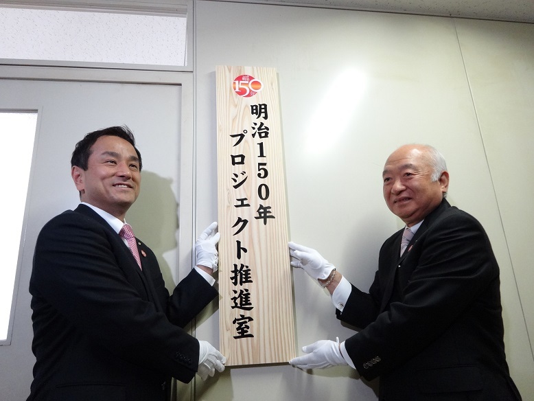 明治150年プロジェクト推進室前で看板の掲出をする柳居議長と村岡知事